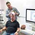A man sits on a chair as a second man stands behind him using an ultrasound device on his head.